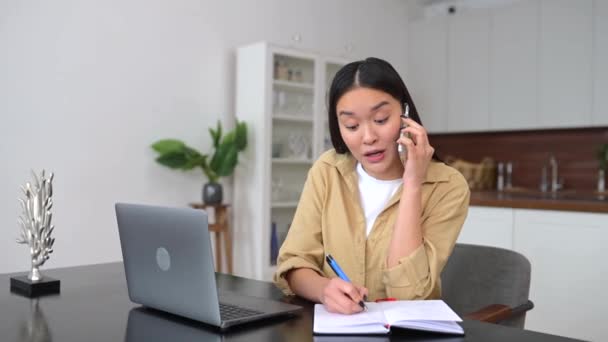 Mulher freelance asiática fala no smartphone e toma notas com caneta — Vídeo de Stock