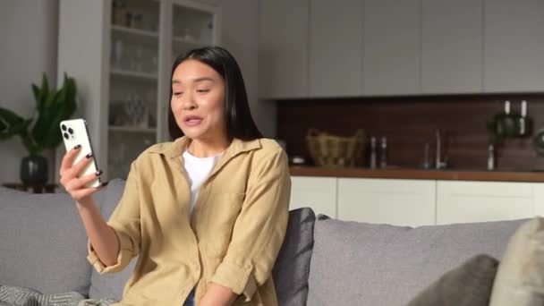 Happy smiling young Asian woman sitting on the sofa talking via video connection — Stock Video