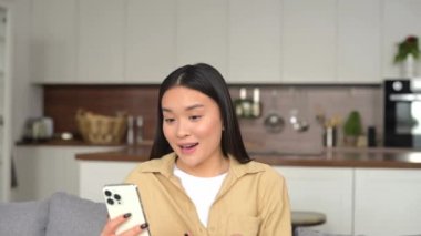 Hilarious asian woman using smartphone for video connection while sitting at the sofa