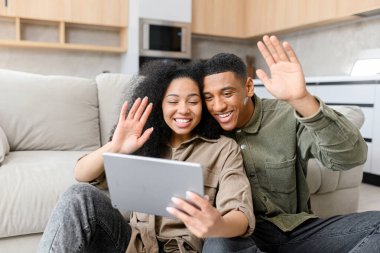 Gündelik giysiler giyen çok ırklı çift tablet ekranına bakıyor ve yerde yan yana otururken el sallıyorlar.