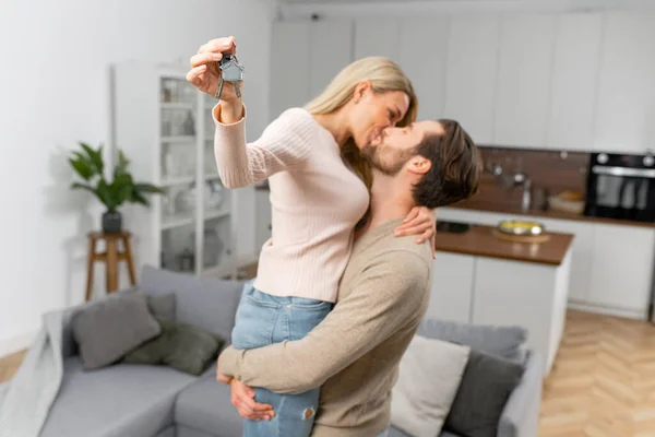 Caucasian woman holding home keys while kissing with her boyfriend in new apartment. Lovely couple feeling ecstatic about their recent purchase of new home — Stock Photo, Image