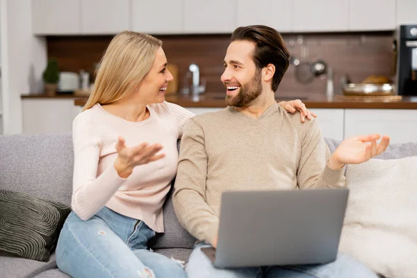 Casal caucasiano sentado no sofá e usando laptop para passar o lazer juntos, assistindo filmes, web surf, compras on-line. Esposos alegres olhando um para o outro — Fotografia de Stock