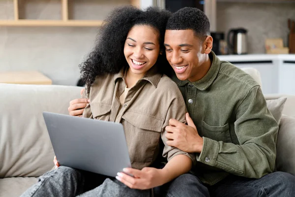 Весела пара закоханих проводить дозвілля онлайн з ноутбуком вдома. Молода жінка і чоловік дивиться на екран ноутбука, сидячи на дивані — стокове фото