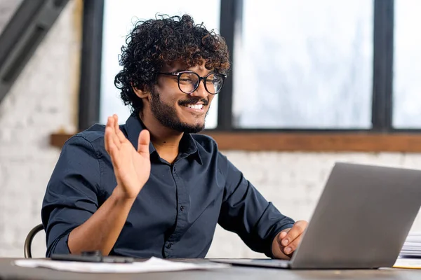 Cheerful attractive Indian guy using laptop for video communication. Guy in glasses sitting at the table in modern office and waving into webcam