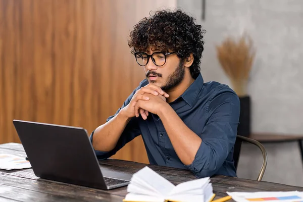 Skupiony i zaniepokojony Indianin w eleganckiej, luźnej koszuli patrzący na ekran laptopa. Poważny facet siedzi przy biurku i spoczywa na podbródku, czując wątpliwości. — Zdjęcie stockowe