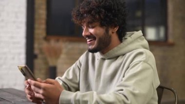 Dahili ofisteki masada oturan, akıllı telefon tutan mutlu, kıvırcık Hintli adam.