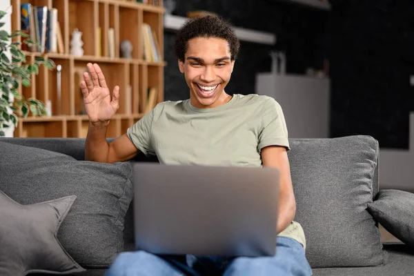 A smiling hipster man talking to his friend or family, waving at the laptop screen — 스톡 사진