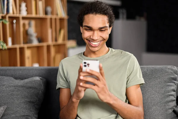 Handsome hipster guy in casual t-shirt sits in comfortable armchair and spends leisure time online — Stock fotografie