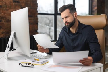 Konsantre Kafkasyalı adam evrak işleriyle uğraşıyor. Ofisinde bilgisayarla masada oturan bir adam kağıtları karıştırıyor. Adam kontrol raporu