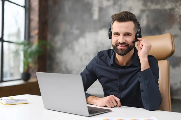 Friendly young guy in smart casual wear is using headset and laptop for online communication, supporting, selling. Guy sitting at the office desk and smiling — Foto de Stock