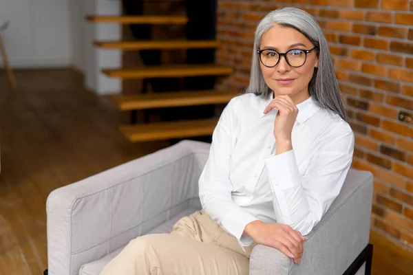 Enchanting carefree middle-aged gray-haired woman sitting at the armchair and relaxing. Portrait of charming mature woman daydreaming at home — Foto de Stock