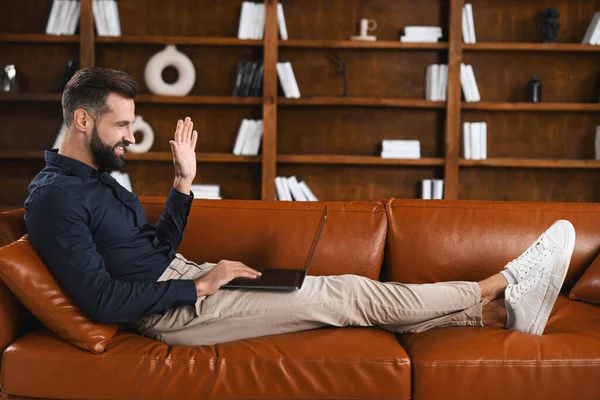 Smiling caucasian man wearing casual wear lying on the sofa and using laptop at modern home office. Confident male entrepreneur waving with hand to the screen with book shelves — Foto Stock