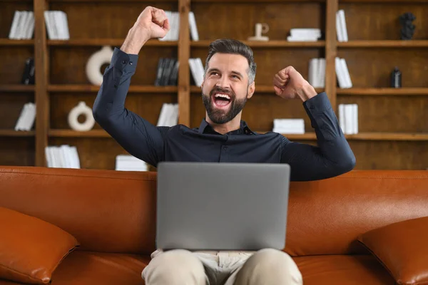 Excited man sitting on the brown sofa looking away in front of the laptop and screaming, surprised emotional lucky man with wide opened mouth — ストック写真