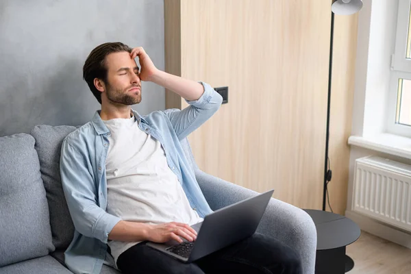 Cansado empreendedor masculino caucasiano sentir dor de cabeça do trabalho on-line com laptop. Homem sério senta-se no sofá e massagear templos — Fotografia de Stock