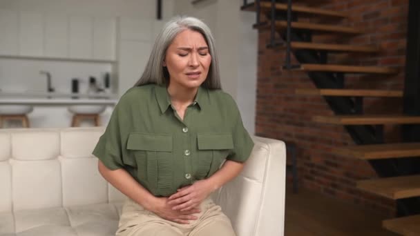 Ανησυχία ώριμη γυναίκα 50 ετών με γκρίζα μαλλιά που πάσχουν από στομαχόπονο κάθεται στον καναπέ — Αρχείο Βίντεο