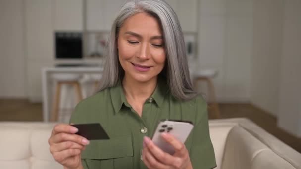 Serene middle-aged grey-haired Asian woman shopping online sitting with credit card and mobile phone — Stockvideo