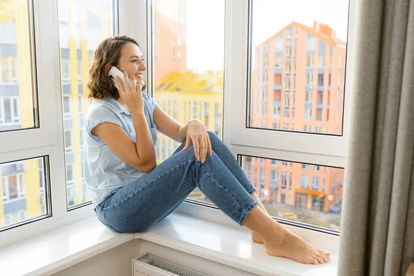 Serene genç bayan evde pencerenin kenarında oturmuş arkadaşlarıyla sohbet ederken akıllı telefondan konuşuyor. Sıradan giyinen çekici bir kadın oturuyor.