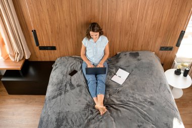 Yatakta oturan ve proje üzerinde çalışan kadın iş kadını ya da kadın serbest çalışanın yüksek açılı görüntüsü.