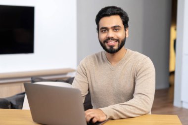 Gündelik kıyafet giyen neşeli Hintli adam masada oturuyor ve modern ofis bilgisayarını kullanıyor.