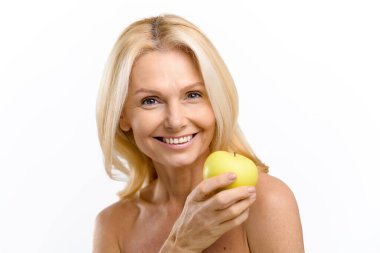 Sağlıklı ve güzel orta yaşlı, çıplak omuzlu, yüzünün yanında yeşil elma tutan bir kadın.