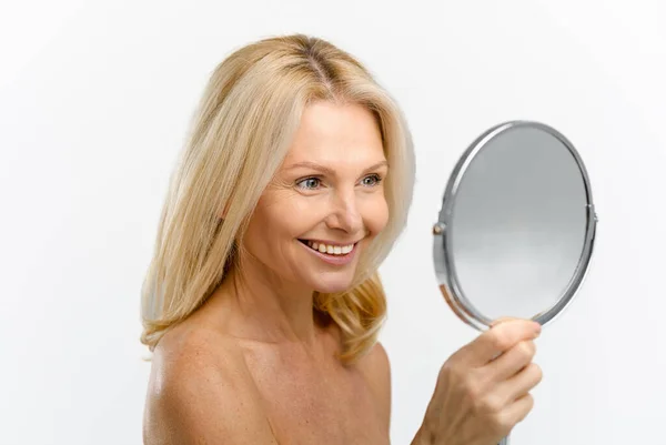 Radosna blondynka po pięćdziesiątce badająca swoje odbicie w lustrze odizolowana — Zdjęcie stockowe