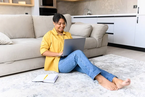 Charming and calm African-American woman sits on the floor in cozy living room with a laptop — стоковое фото