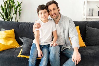 Keyifli baba-oğul portresi rahat bir oturma odasında kanepede oturuyor, kameraya bakıyor ve gülüyor, hafta sonunun keyfini çıkarıyor.