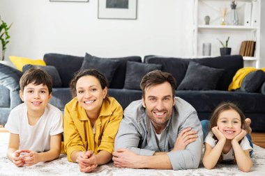 Dört kişilik neşeli, çok ırklı bir ailenin portresi. Anne, baba, oğul ve küçük kız, rahat oturma odasında yere uzanmış kameraya bakıyor ve gülüyor.