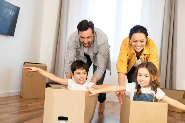 Многорасовая семья из четырех человек веселится в новом доме, родители катают детей в картонных коробках в пустой гостиной, счастливые дети размахивают руками и смеются — стоковое фото