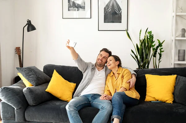 Happy caucasian couple of spouses sitting at the couch at home and smartphone. Man and woman making selfie — Fotografia de Stock