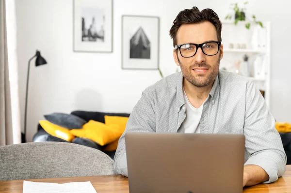 Handsome young freelancer man with beard in glasses sitting at the table and looking at the camera, working on laptop — Zdjęcie stockowe