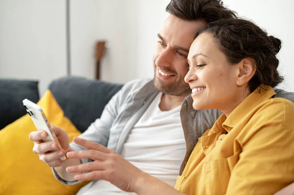 Happy caucasian couple sitting at the couch at home and holding smartphone. Man and woman watching movie — Stockfoto