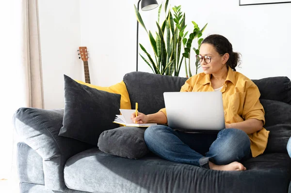 O femeie independentă concentrată cercetează sarcinile de afaceri, luând notițe cu un laptop acasă. Femeia scrie într-un notebook, se uită la cursuri online, webinar — Fotografie, imagine de stoc