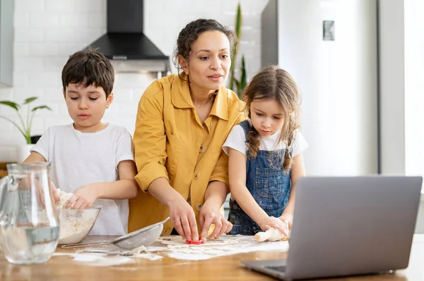 Middle-aged mother and her kids cooking together, kneads the dough, using rolling pin. Mom and kids spends leisure at home, baking cookies, looking tutorial at the laptop — Photo