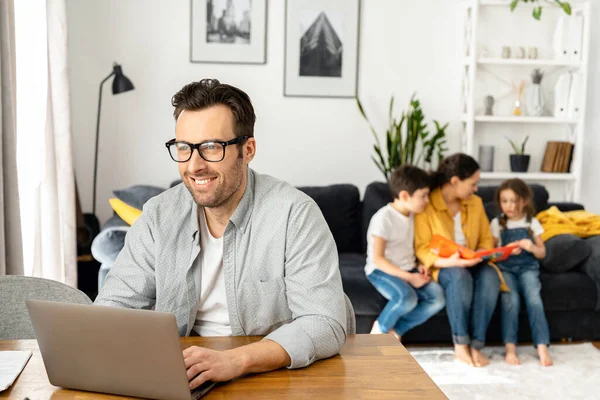 O pai feliz a usar óculos está a trabalhar à distância. Homem usando laptop para trabalho freelance sentado à mesa em casa, enquanto a família sentada pacificamente juntos — Fotografia de Stock