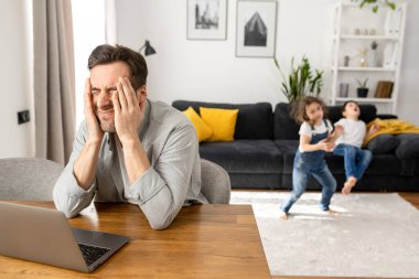 Canı sıkkın baba dizüstü bilgisayarı kapalı oturuyor, iki çocuğu zıplarken ve bağırırken kafasını ellerle kapatıyor.