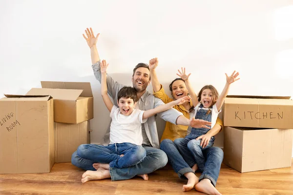 Happy multiracial family of four sitting on floor surrounded cardboard boxes in empty living room. Owners of estate — Stock Photo, Image