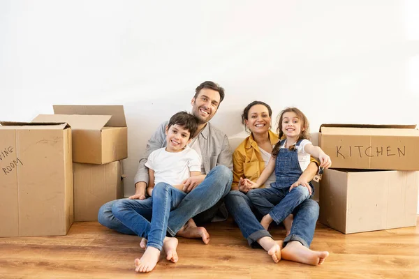 Cute daughter holding keys with keychain in form of little house. Overjoyed multiracial family of four sitting on floor surrounded cardboard boxes in empty living room — Stock Photo, Image