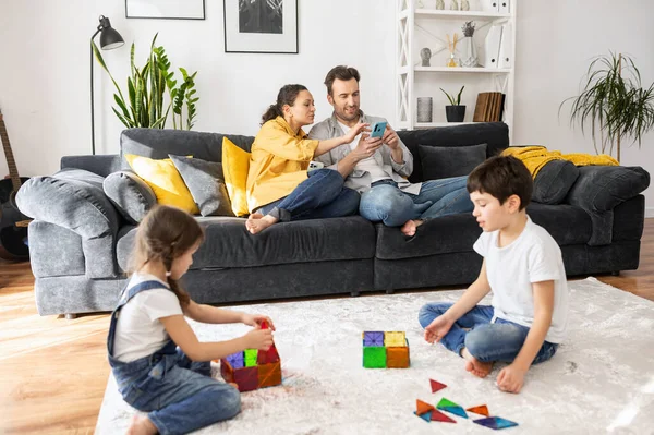 两个家长在舒适的客厅里，用智能手机坐在沙发上，一边翻看新闻，一边两个孩子和建筑商一起在地板上玩耍，兄弟们一起享受着游戏 — 图库照片