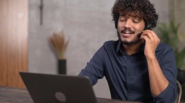 Kablosuz kulaklık takan gülümseyen etnik Hintli adam sanal bağlantı için laptop kullanıyor.