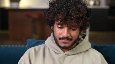 Kanepede otururken dizüstü bilgisayar kullanan gündelik giyinen iyimser etnik bir adam.