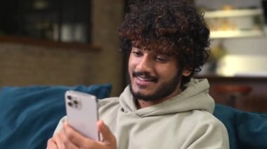 Focused bearded Indian man sitting on sofa at home and holding smartphone