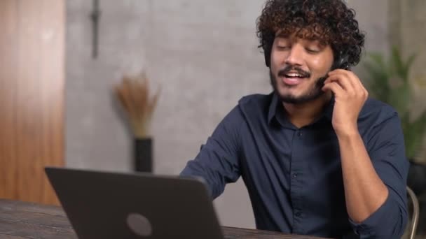 Sonriente hombre indio étnico que usa auriculares inalámbricos con computadora portátil para la conexión virtual — Vídeos de Stock