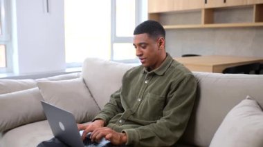 Normal giyinen iyimser adam kanepede otururken dizüstü bilgisayar kullanıyor.