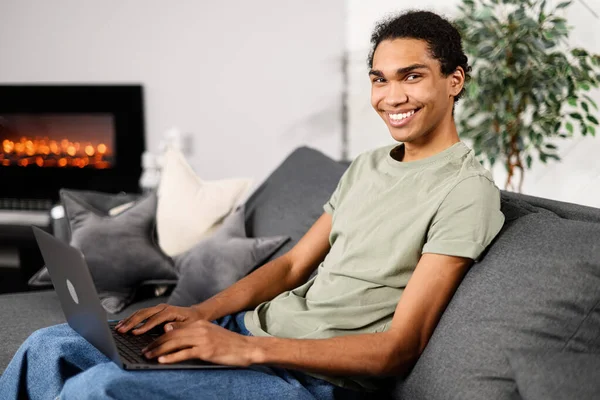 Kanepede oturan ve modern merkez ofisteki dizüstü bilgisayar kullanan, gündelik giyinen neşeli, çok ırklı adamın yan görüntüsü. İyi ki erkek girişimci klavyede yazıyor.