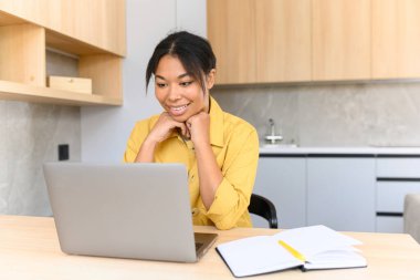 Neşeli, melez bir kız evde mutfaktayken bilgisayarını uzaktan çalışmak ya da boş vakit geçirmek için kullanır. Yan manzara, afro saçlı güzel bir kız ekrana bakıyor.