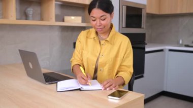 Mutfakta dizüstü bilgisayarla masada oturan, gülümseyen, çok ırklı genç bayan girişimci.