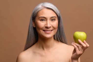 Beyaz saçlı, orta yaşlı, çıplak omuzlu, elma tutan güzel bir Asyalı kadın.