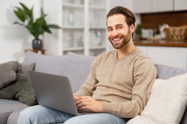 Gülümseyen genç sakallı adam evde dizüstü bilgisayar kullanıyor. Kanepede oturan, internette sörf yapan, haberleri izleyen bir adam.