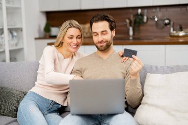 Neşeli beyaz çift bilgisayar ekranına bakıyor ve online işlem yapmak için kredi kartı numaralarını kontrol ediyor. Gülümseyen eşler online alışveriş yapıyor. Para aktarımı konsepti
